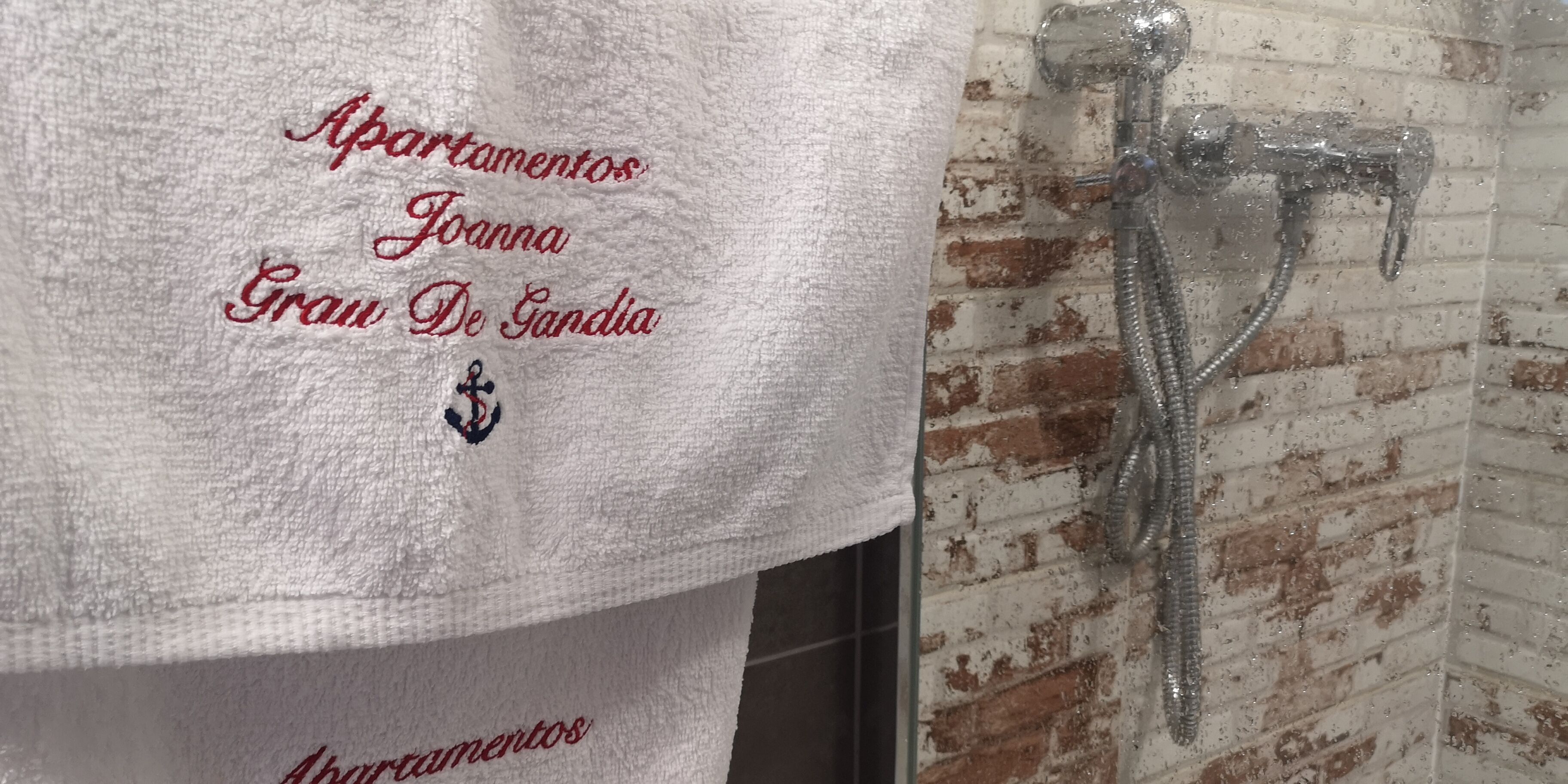 Een witte handdoek met geborduurd tekst en een logo, hangend aan een bakstenen muur nabij een douche.