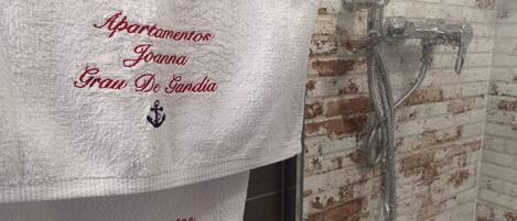 Une serviette blanche ornée d’un texte brodé et d’un logo, accrochée à un mur de briques près d’une douche.