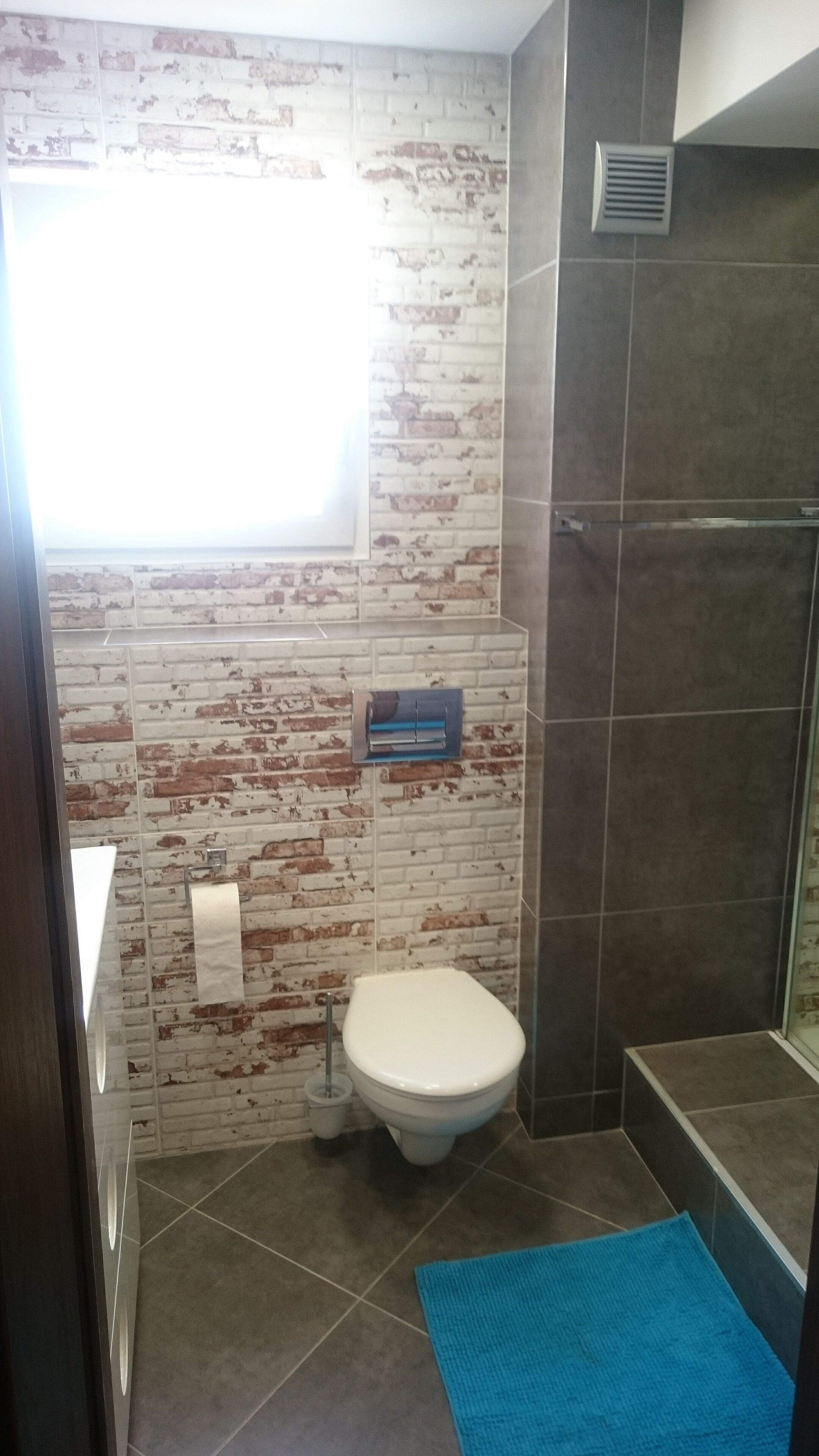 Een badkamer met een wit toilet, een blauwe mat en een muur met een baksteenmotief.