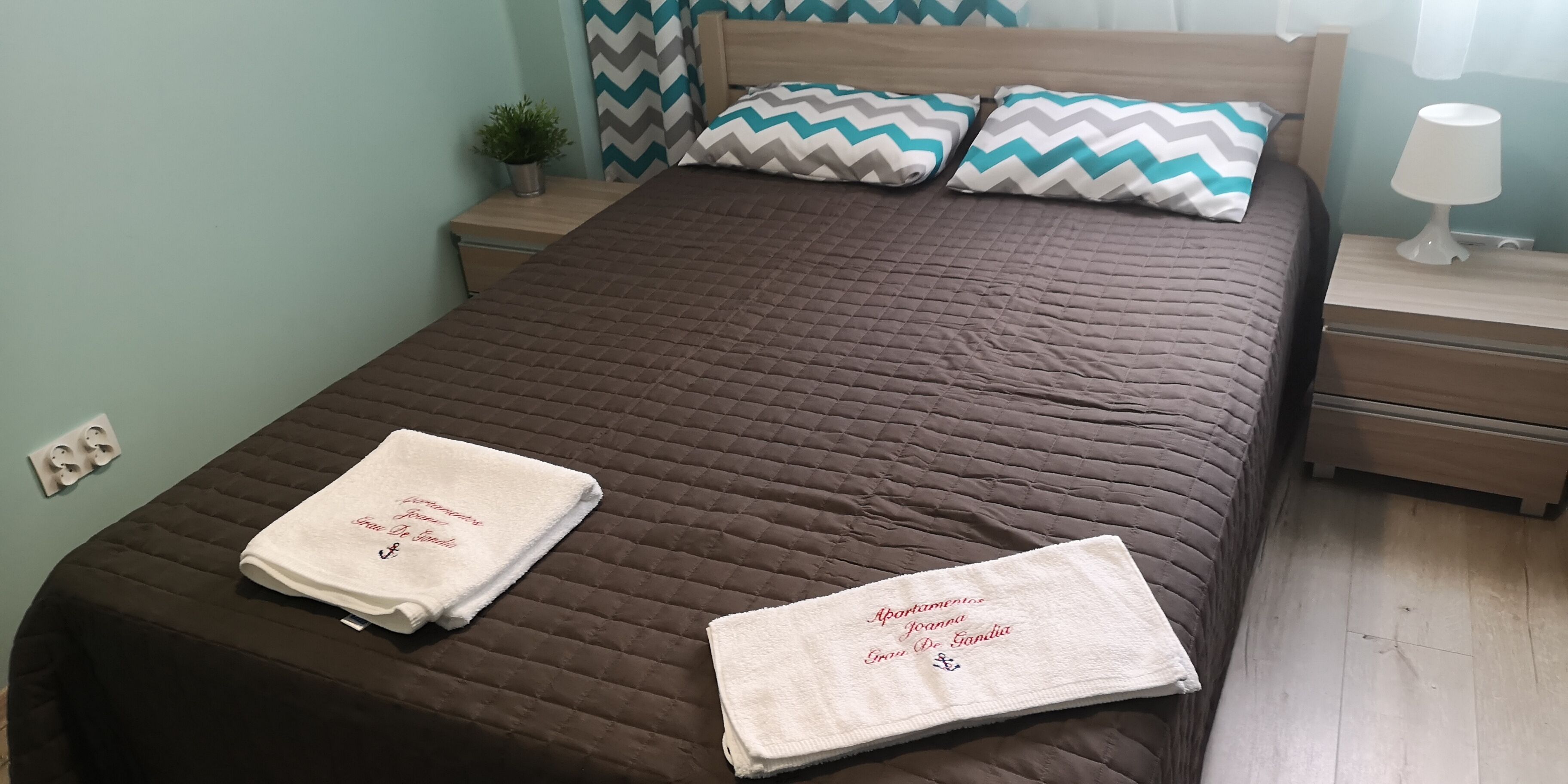 Una cama bien hecha con dos toallas, una mesita de noche con lámpara y un enchufe.