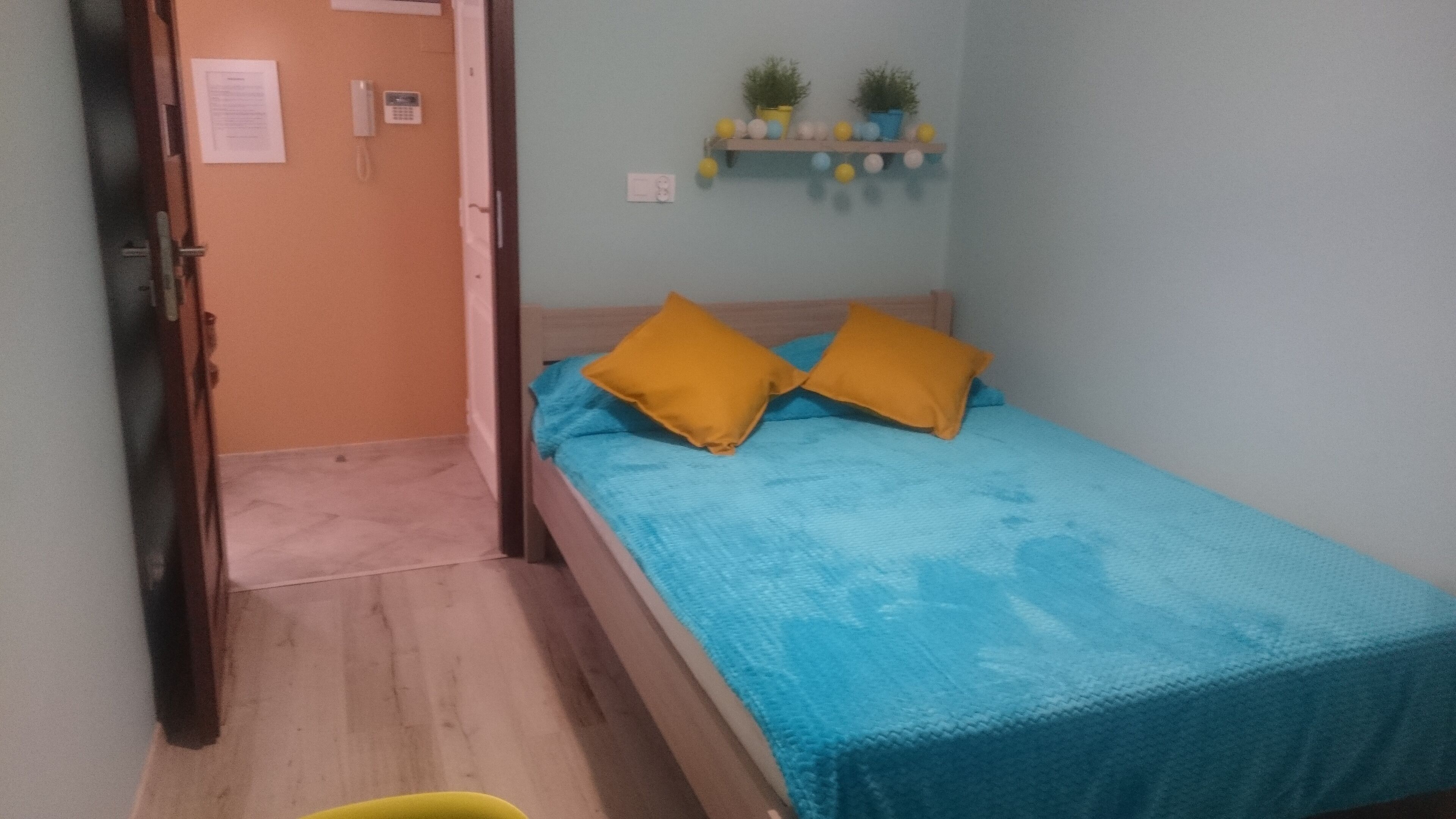 Une petite chambre avec un lit simple, une couvre-lit bleue, deux oreillers jaunes, une porte en bois, une petite étagère avec des plantes et un interrupteur sur le mur.