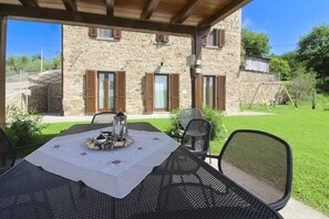 Une terrasse extérieure avec une table dressée pour deux personnes, un bâtiment en pierre avec des volets en bois, et une balançoire en arrière-plan.