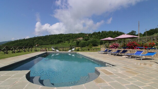 Una piscina rectangular con solárium de piedra, rodeada de sillas de descanso y sombrillas, con colinas verdes y cielo despejado al fondo.
