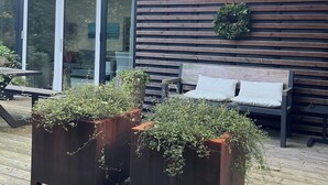 Une terrasse extérieure moderne avec un revêtement de sol en bois, une enceinte en verre et un mur en bois avec des lattes horizontales. On y trouve deux jardinières en bois ornées de plantes et un banc avec des coussins.