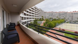 Un balcón con mobiliario, vista a un patio con vegetación y una cancha de tenis, y un edificio de varios pisos al fondo.
