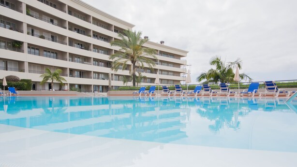 Zona de piscina de hotel con numerosas tumbonas y un edificio grande al fondo.