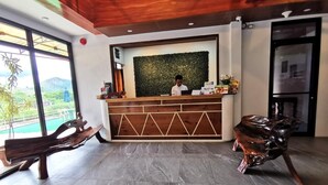 A reception area with a wooden reception desk, a green wall, and a view of a mountain and pool. - Charms Hotel (Coron)
