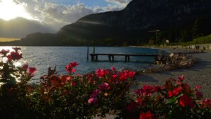 Vista del lago con un muelle que se adentra en el agua, rodeado de flores rojas y montañas al fondo.