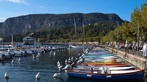 Una marina con numerosos barcos amarrados a lo largo de la costa, una montaña al fondo y un paseo marítimo con gente caminando.