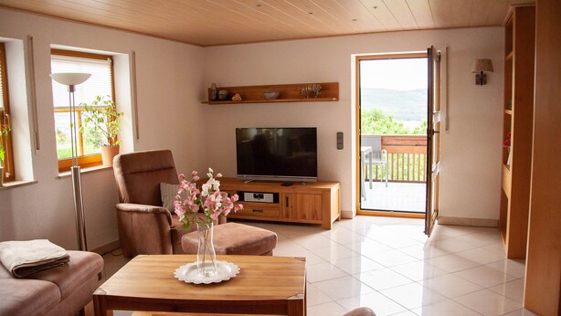 Una sala de estar con televisor, muebles de madera, una planta en maceta y vistas a través de un ventanal.