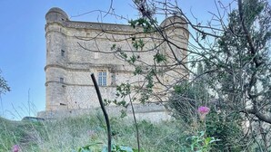 A stone building with a cylindrical tower, surrounded by lush greenery and flowering plants. - Vaucluse- Pied du Ventoux- Le Barroux- Gravel house (Le Barroux)