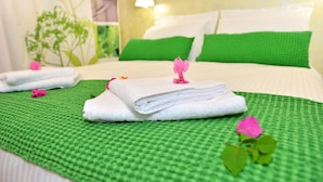 Una cama bien hecha con ropa de cama verde y blanca, un tapete texturizado verde y una flor rosa sobre los almohadones.