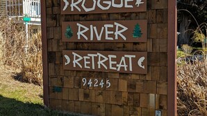 A wooden sign for Rogue River Retreat with a green roof, surrounded by grass and trees. - Where the river meets the beautiful Oregon Coastline. (Gold Beach)