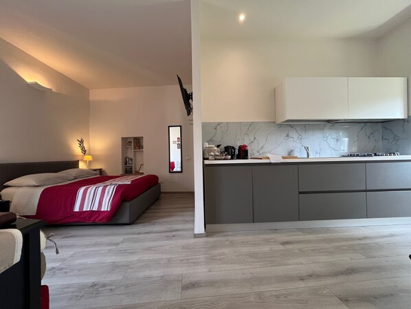 A modern bedroom with a bed, bedside table, and a nightstand. Adjacent to the bedroom is a kitchen with a countertop, cabinets, and a stove.