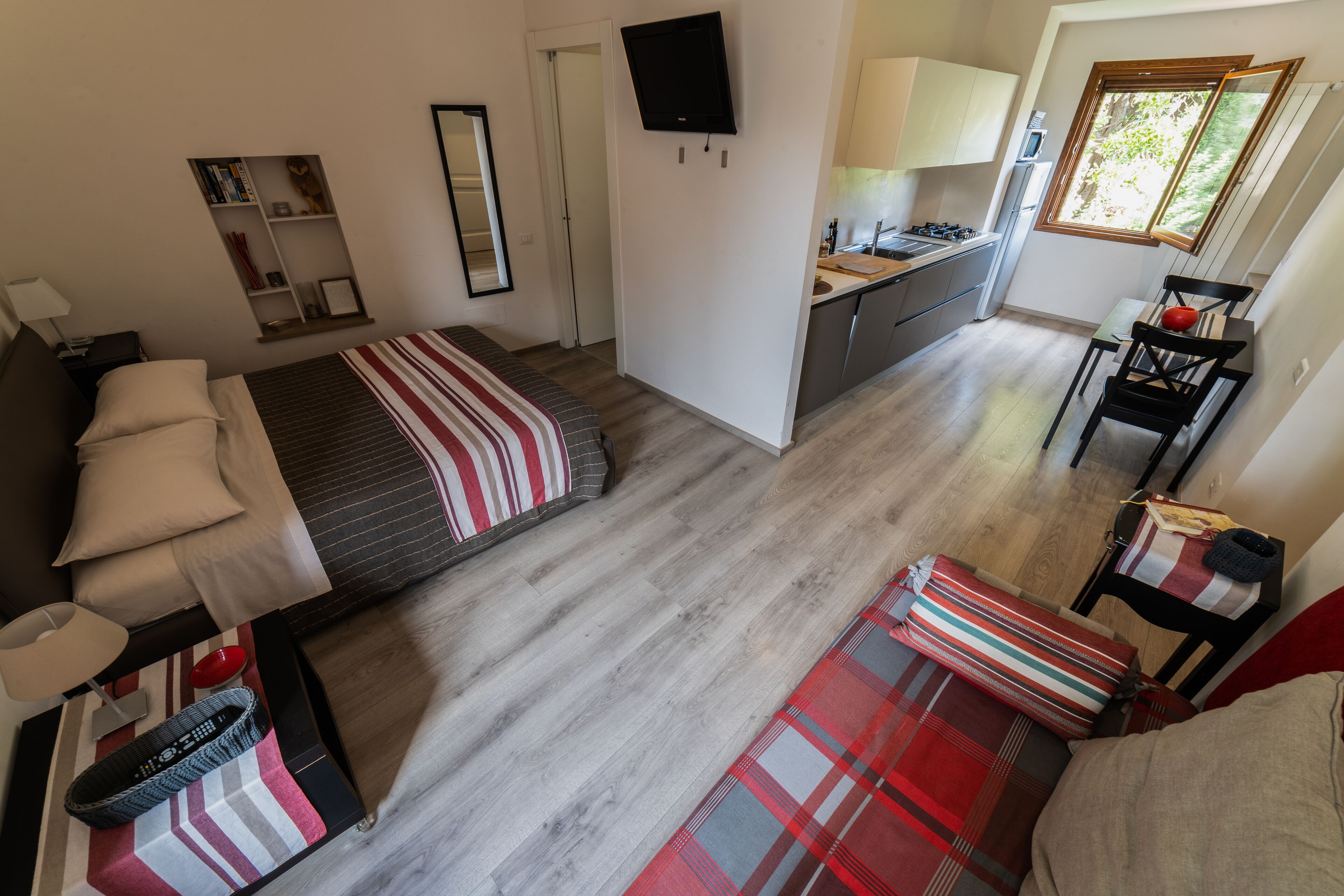 A compact hotel room with a bed, bedside table, lamp, and a small seating area with a plaid blanket.