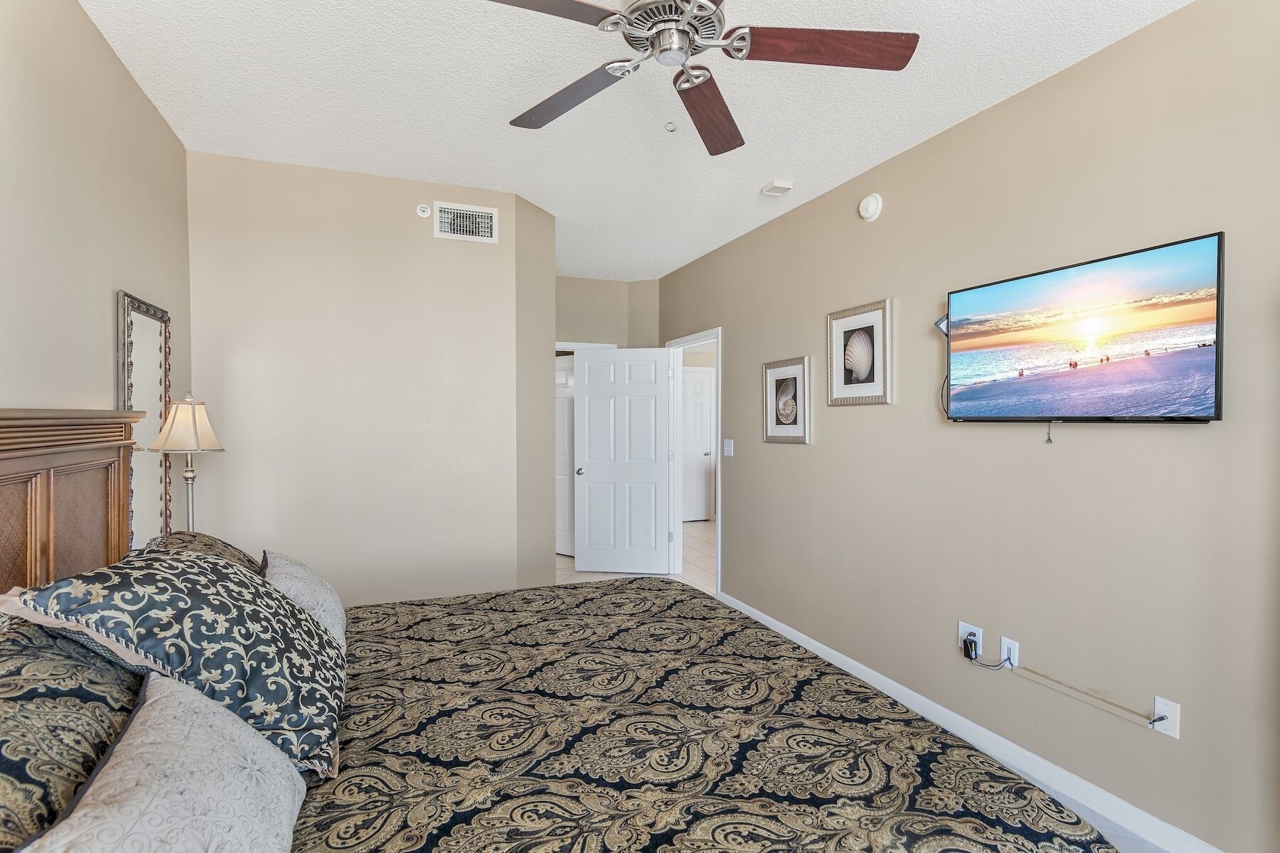 A bedroom with a patterned carpet, a ceiling fan, a television displaying a beach scene, and a door leading to another room.