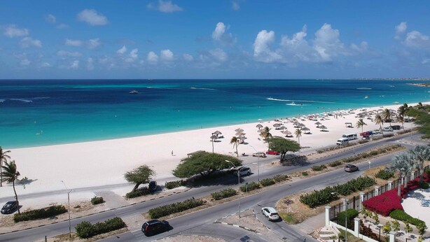 Een strand met wit zand, helder turquoise water en een weg met geparkeerde auto's.