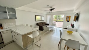 Een moderne keuken met een marmeren aanrecht en witte kasten, een eethoek met een witte tafel en stoelen, en een zithoek met een bank en een plafondventilator.