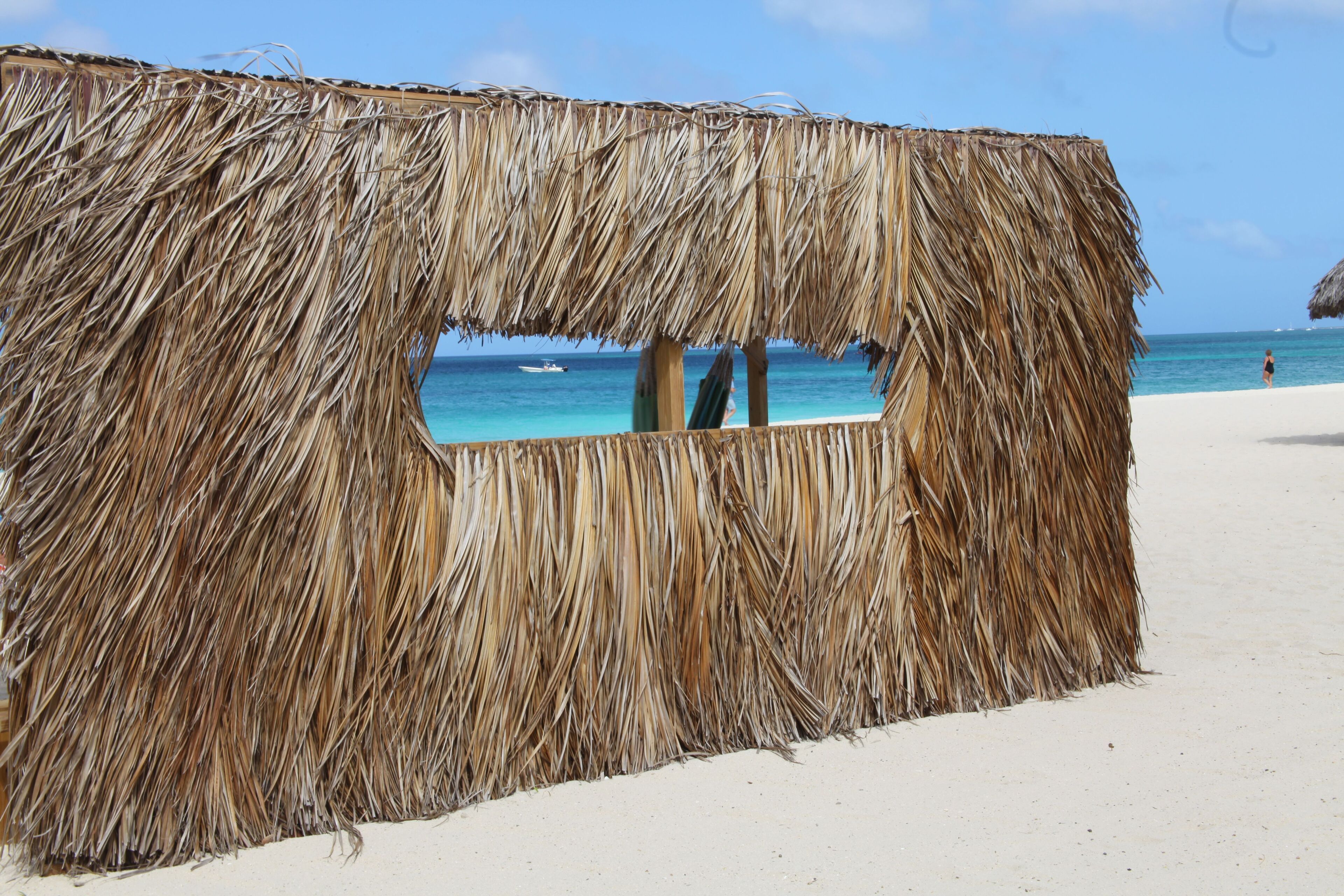 Une construction à toit de chaume, dotée d’une fenêtre, qui offre une vue sur une plage aux eaux bleu clair et sur un bateau au loin.