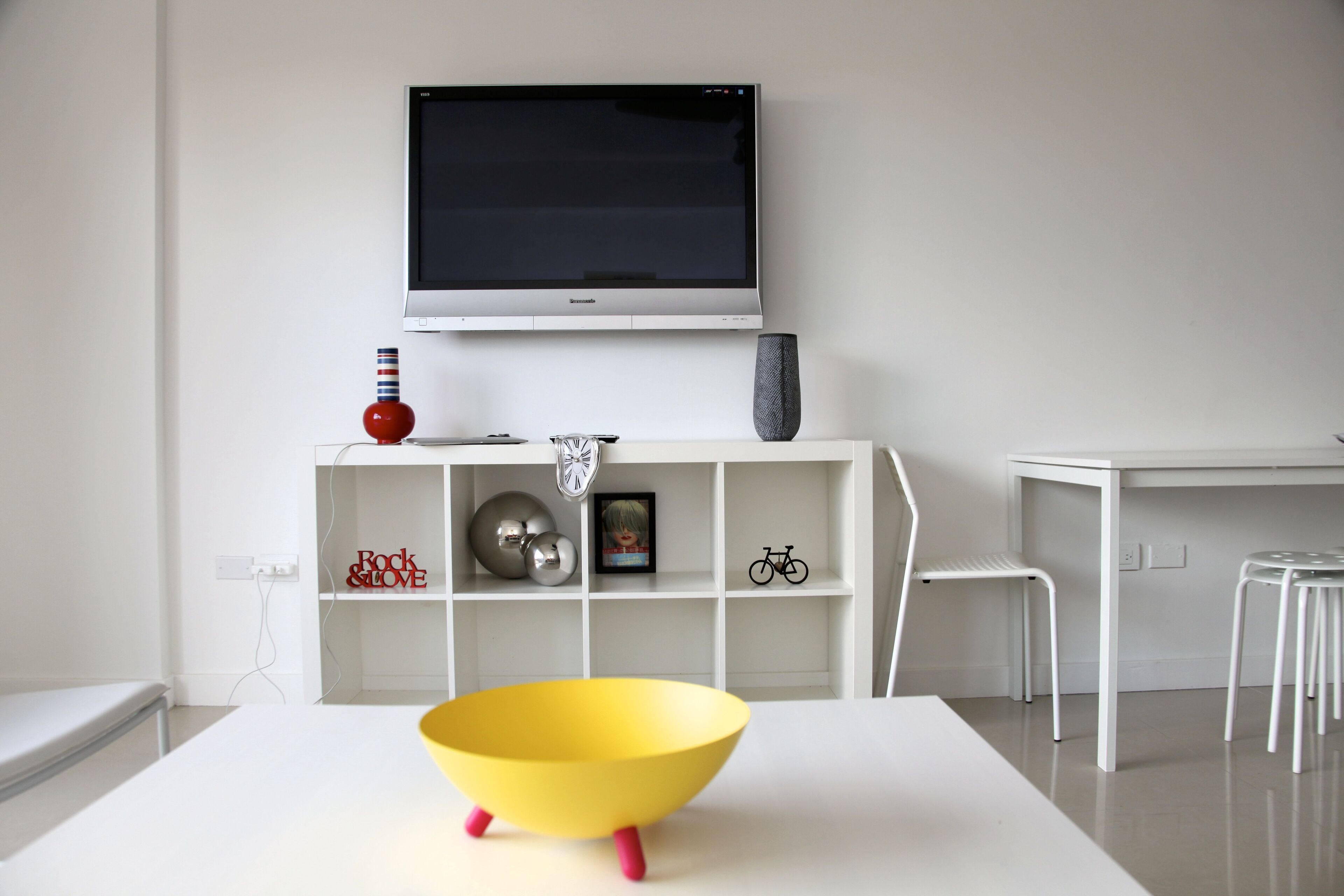 A modern living room with a flat-screen TV mounted on the wall, a white console table with decorative items, a yellow bowl on a white table, and white chairs.