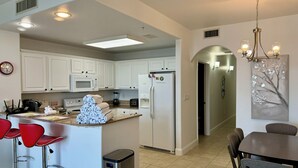 A modern kitchen with white cabinets, a refrigerator, a microwave, and a coffee machine. There are red bar stools and a dining table with chairs. - New! 1-King Bedroom, 2 baths with Breathtaking view at Blue Heron Beach Resort (Orlando)