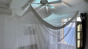 Una habitación con una cama bajo una mosquitera, un ventilador de techo y una ventana con cortinas.