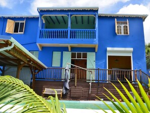 A two-story building with a blue exterior, a balcony on the upper floor, and a wooden deck in front. - Villa with exceptional sea view, infinity pool and jacuzzi, direct sea access (POINTE NOIRE)
