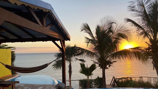 Una piscina con vistas a la puesta de sol sobre el océano, rodeada de palmeras y una terraza de madera.