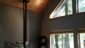 A cozy living room with a stone fireplace, a bookshelf, and a large window offering a view of trees. - Greenwater chalet/cabin - 3 bed + loft - 2 full baths (Enumclaw)