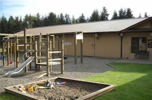 A playground with a slide and climbing structures, a sandbox, and a building with a metal roof.