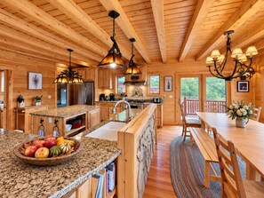 Interior de una cabaña de madera con cocina, comedor y vistas al exterior.