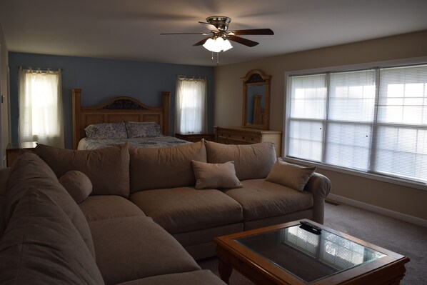 A living room with a sectional sofa, a glass coffee table, a ceiling fan, a bed with a wooden headboard, and a large window with blinds.