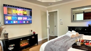 A modern hotel room with a flat-screen TV displaying a movie, a fireplace, a bed with a fluffy blanket, and a tray with towels and a drink. - Premier Secluded Oasis w/ Diving Pool, Hot Tub, Koi Pond & Outdoor Shower (Plano)