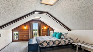 Ein Schlafzimmer im Dachgeschoss mit geneigter Decke, einem Bett mit gemusterten Textilien, einer Sitzbank, einem kleinen Tisch und einem Fenster mit Vorhängen.