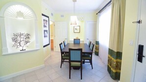 A dining room with a large table, chairs, and a chandelier. There are framed pictures on the walls and a window with a tree decoration.