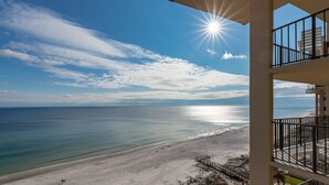 A beachfront hotel with a balcony overlooking the ocean, a clear sky, and the sun shining.