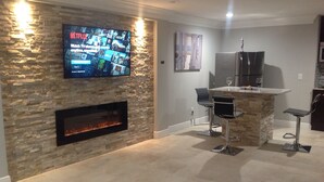 A modern living room with a stone fireplace, a flat-screen TV mounted on the wall, and a kitchen area with a stainless steel refrigerator and a bar counter.