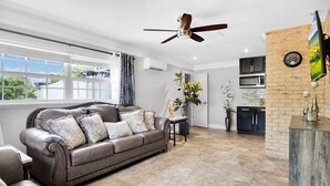 A spacious living area with a leather sofa, a dining table, and a kitchen area with a brick wall and a clock.