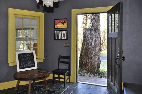 Una habitación con una puerta amarilla, una silla negra, una mesa pequeña con un cartel de pizarra y cuadros en la pared.