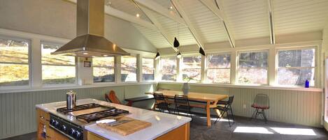 A kitchen with a central island, wooden cabinets, a stove, and a large window overlooking a natural landscape.