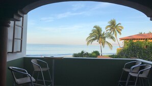 A balcony with two metal chairs and a table, overlooking a beach with palm trees and a building with a red roof. - A pool and a beach… What else do you need? Check out my REVIEWS! GREEN! (BUCERIAS)
