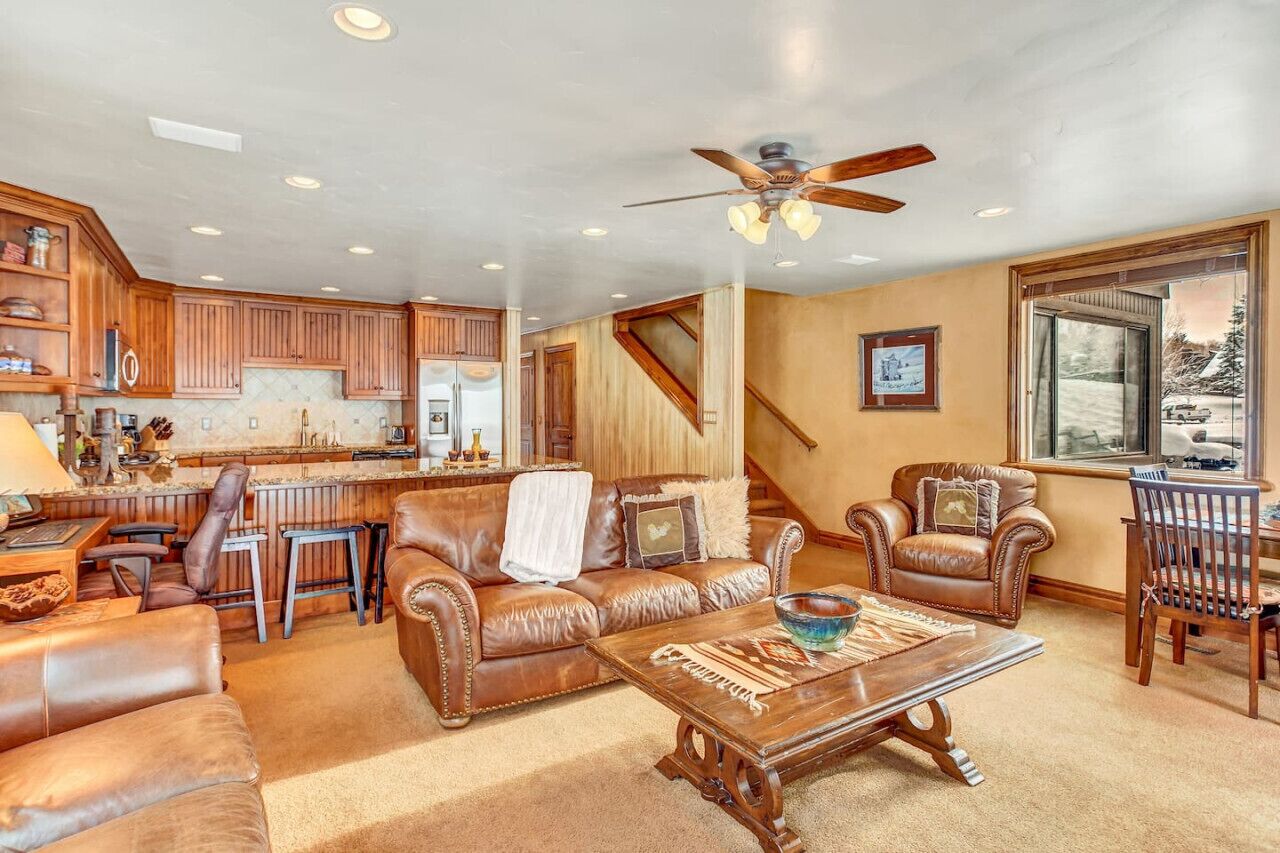 A spacious living area with a leather sofa, coffee table, and a dining table. A kitchen is visible in the background with wooden cabinets and a bar area.