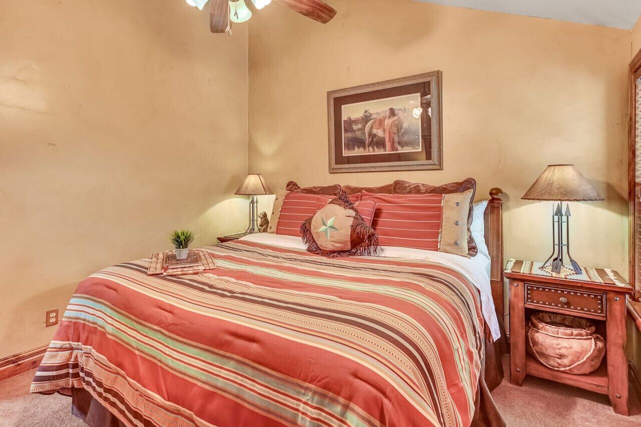 Quarto com cama, duas mesas de cabeceira, um criado-mudo, abajur, quadro na parede e uma pequena planta na cama.