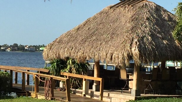 Une construction à toit de chaume, dotée d’une terrasse en bois et de chaises, qui offre une vue sur une étendue d’eau.