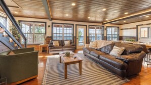 A spacious living room with a wooden ceiling, large windows, a brown leather sofa, a green armchair, and a wooden coffee table. - Mountain High Cabin with hot tub, fire pit, & indoor pool! (Bryant)