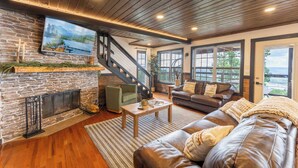 A cozy living room with a stone fireplace, leather sofa, and a staircase leading to another level. - Mountain High Cabin with hot tub, fire pit, & indoor pool! (Bryant)