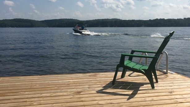 A wooden deck with a green chair and a bench, overlooking a lake with a boat in the distance. - Muskoka, Cozy 2 Bedroom / 2 Bathroom Cottage located on the coveted Lake of Bay (Dwight)