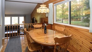 A dining area with a round wooden table, chairs, and a chandelier. A window with a view of trees is visible. - Muskoka, Cozy 2 Bedroom / 2 Bathroom Cottage located on the coveted Lake of Bay (Dwight)