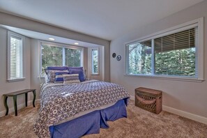 A bedroom with a large bed, two windows with blinds, a small bench, and a wicker basket.