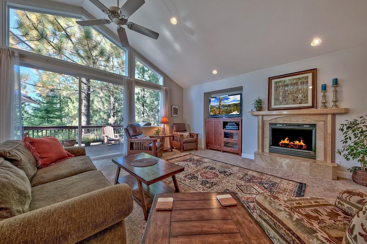 A spacious living room with a fireplace, large windows, and a ceiling fan.
